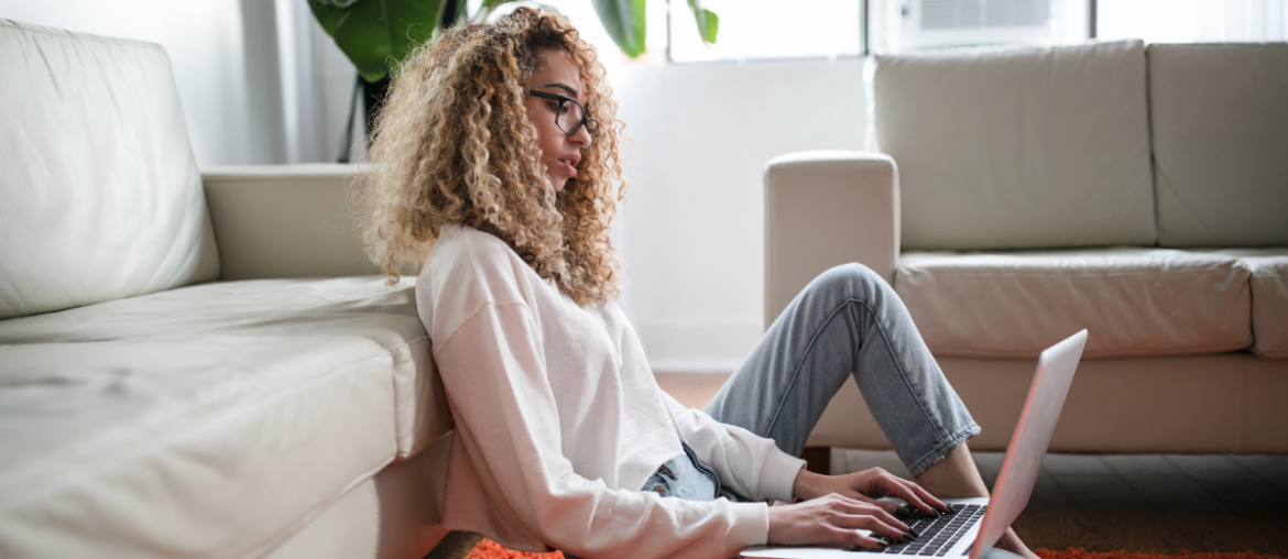 A woman sitting on the floor, leaned against her sofa with her laptop on her lap.